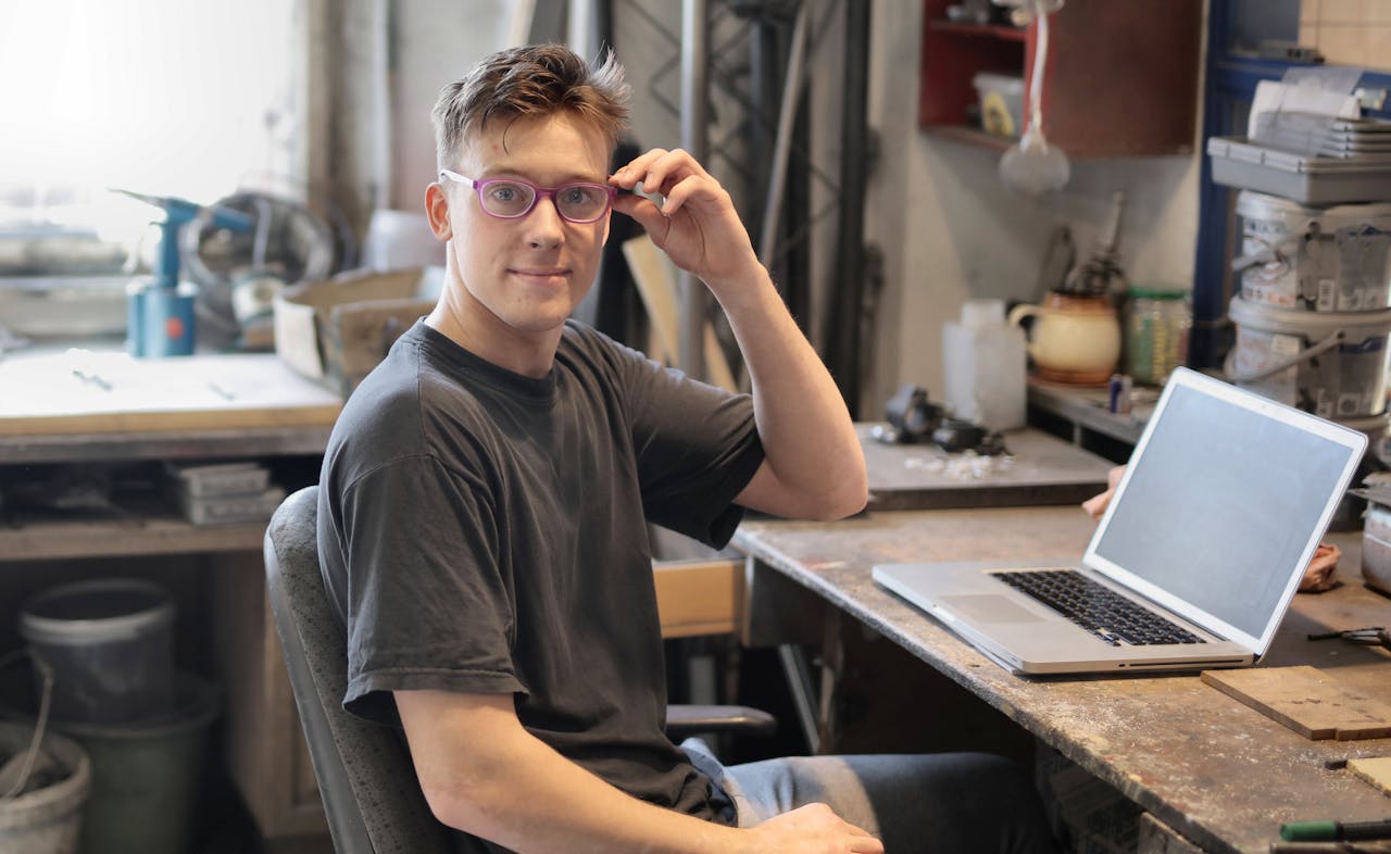 Young man in glasses working remotely on laptop in a workshop setting.