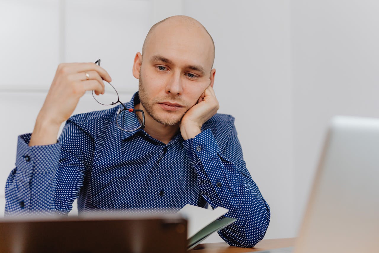 Bald businessman in blue shirt thinking at desk with glasses and laptop.